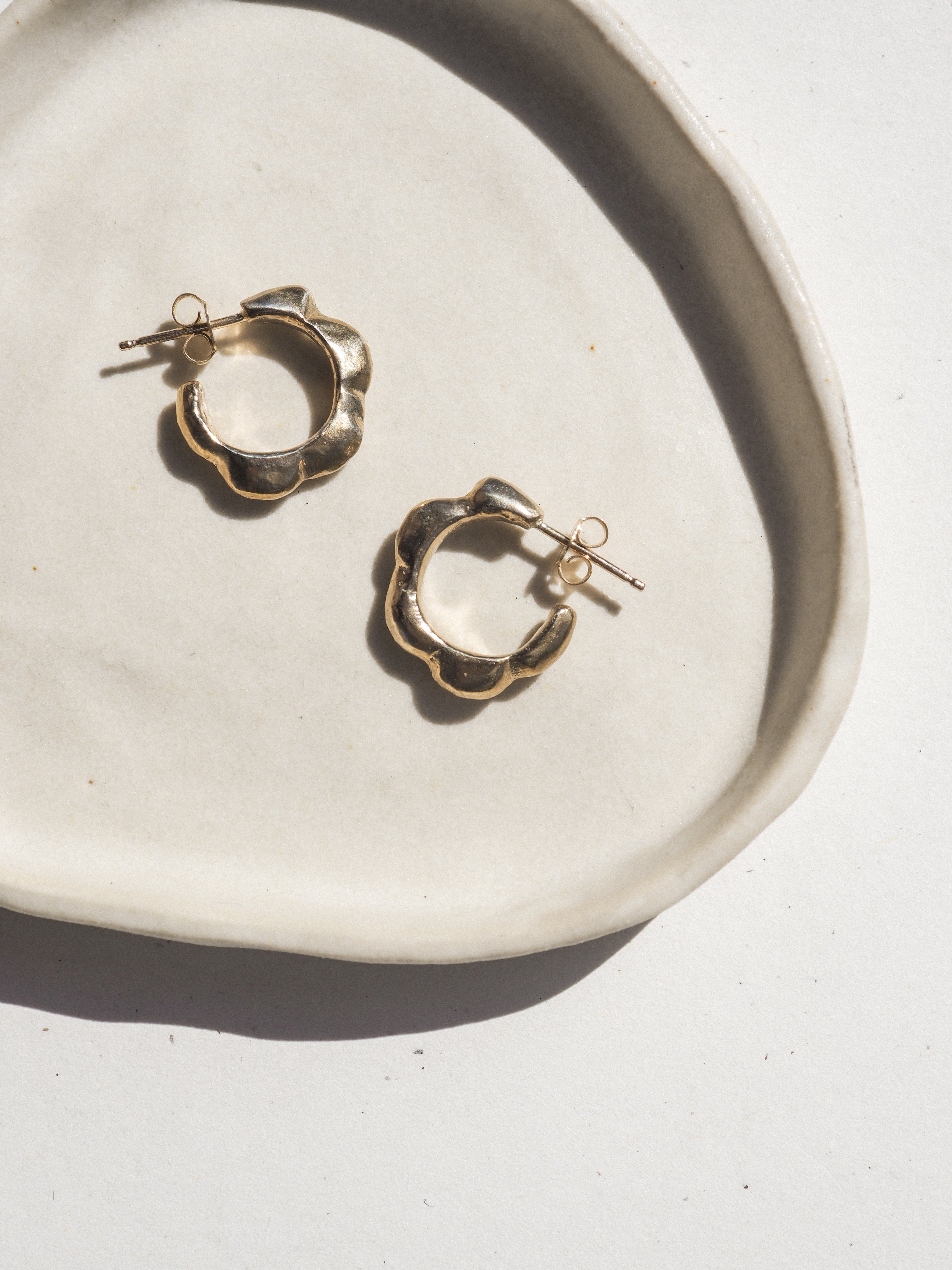 A pair of gold-toned textured hoop earrings displayed on a white ceramic dish against a white background with strong natural shadows.