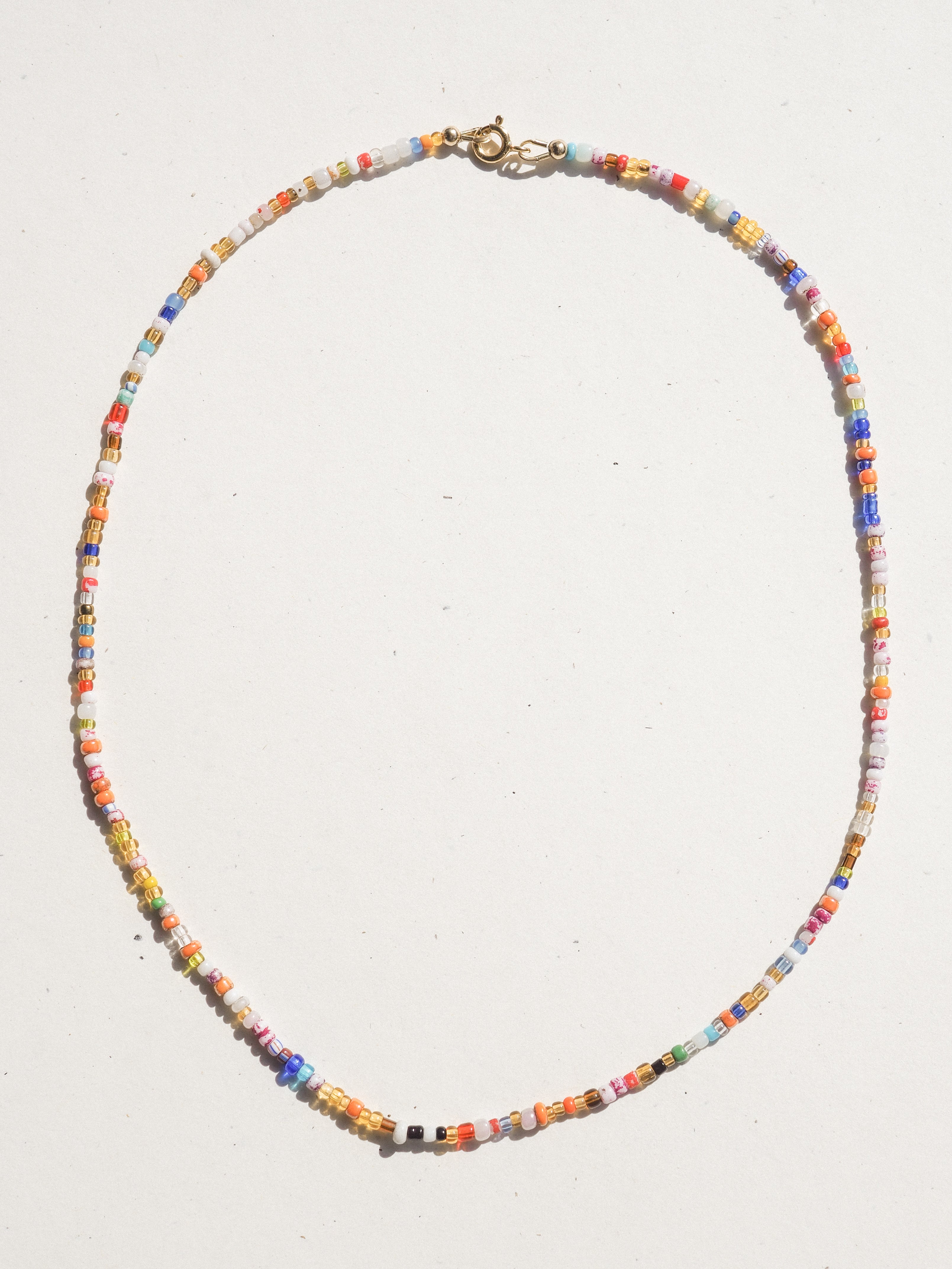 A delicate beaded necklace made of small multicolored cylindrical beads, arranged in an oval shape on a plain off-white background. It features a gold-toned lobster clasp at the top.