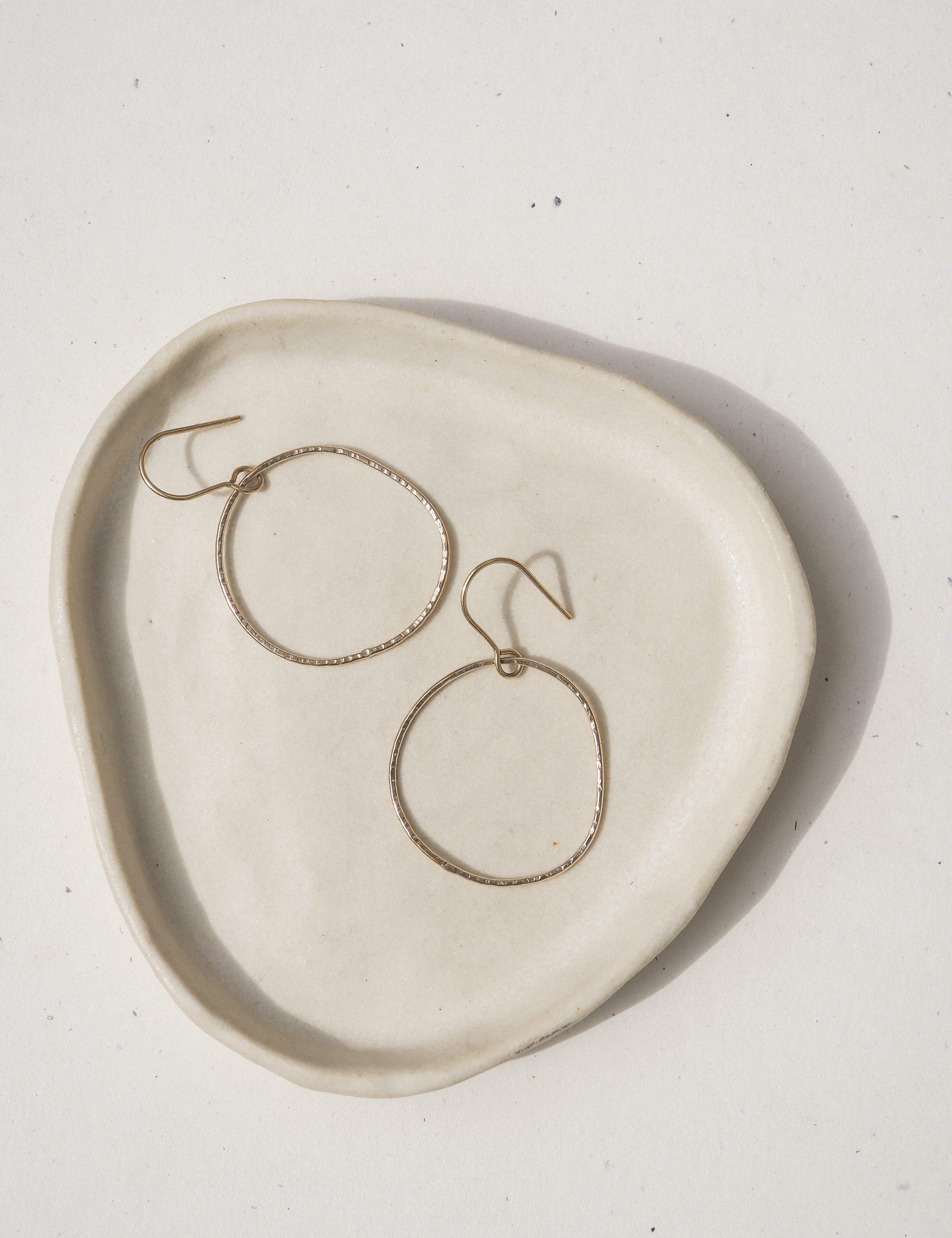 A pair of textured gold-tone hoop earrings resting on an organic-shaped, off-white ceramic dish against a light beige background.