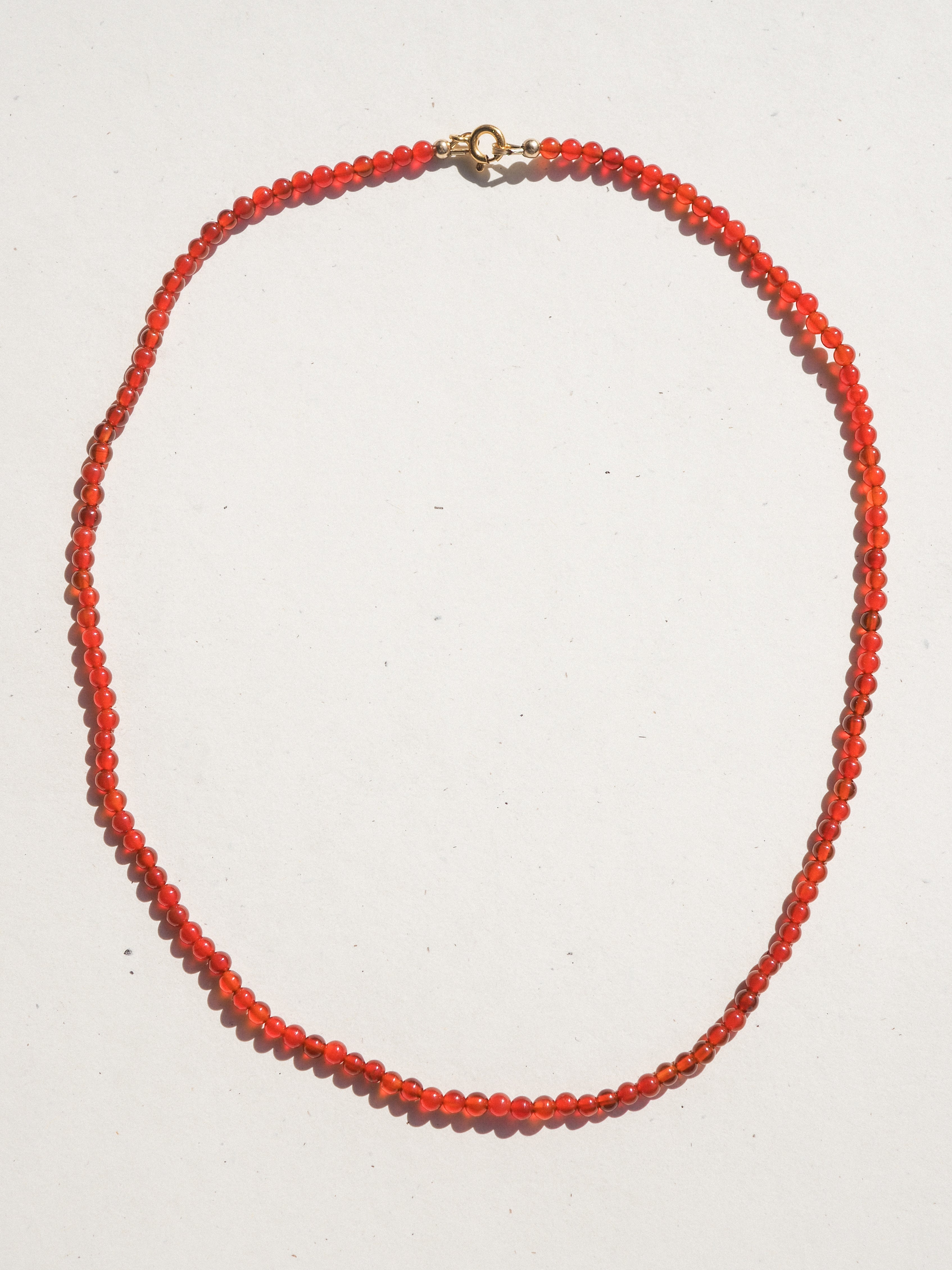 A beaded necklace made of small, faceted orange-red beads with a gold clasp, laid out in an oval shape on a plain white background.