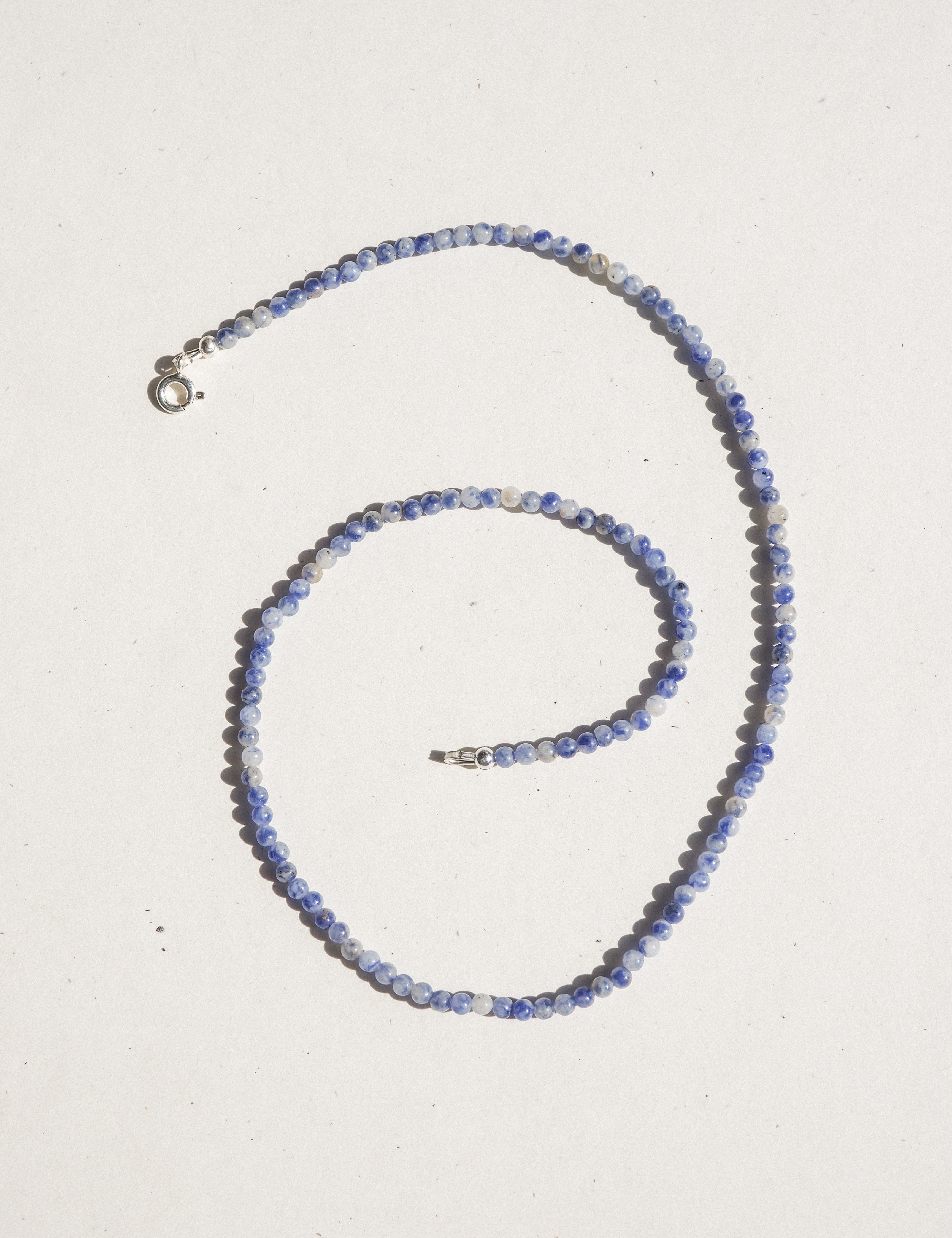A beaded necklace made of small blue and white marbled beads, arranged in a spiral on a light off-white background. The necklace features silver-toned metal clasp hardware.