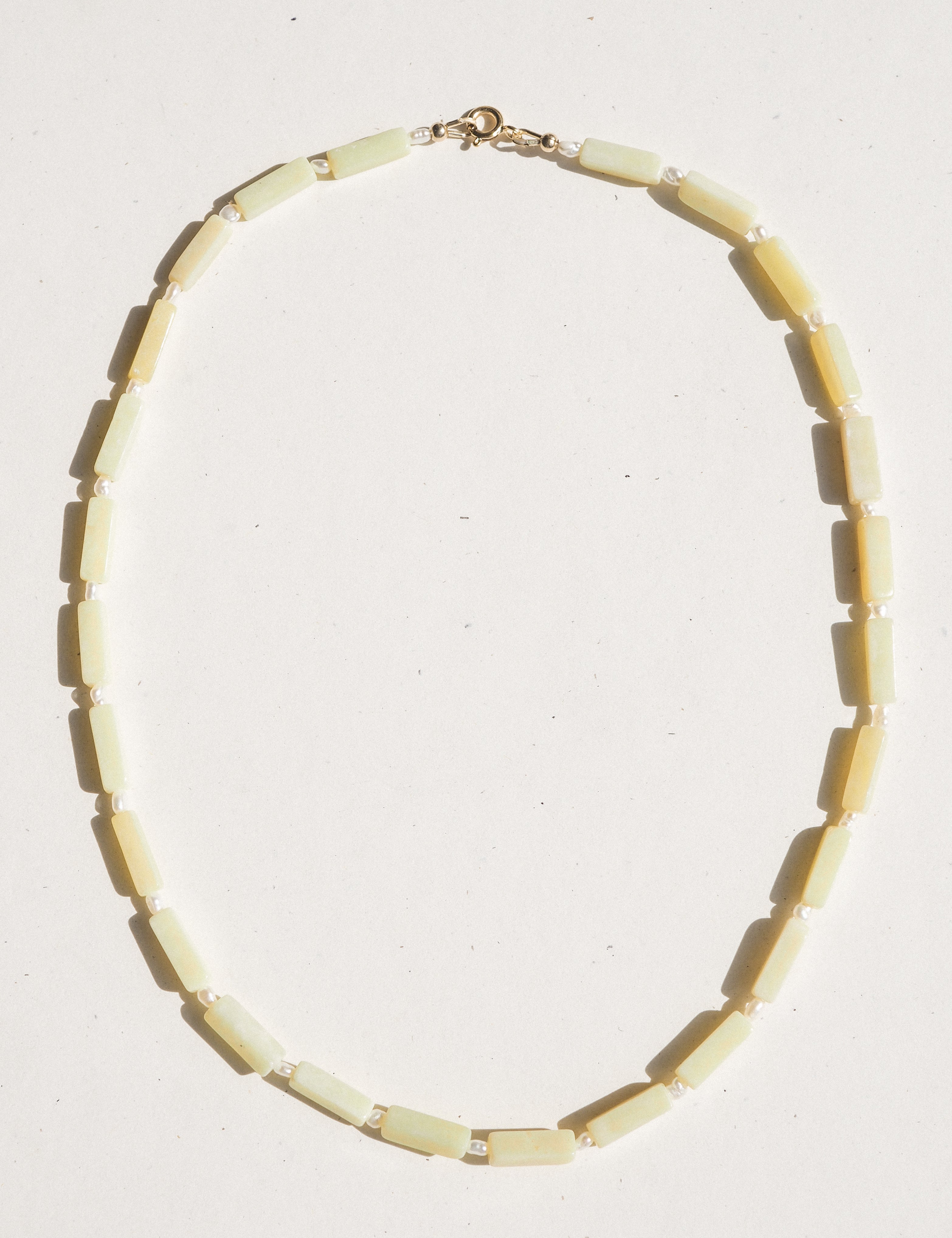 A beaded necklace made of rectangular pale yellow beads separated by small freshwater pearl beads, with a gold clasp, laid flat on a white background.