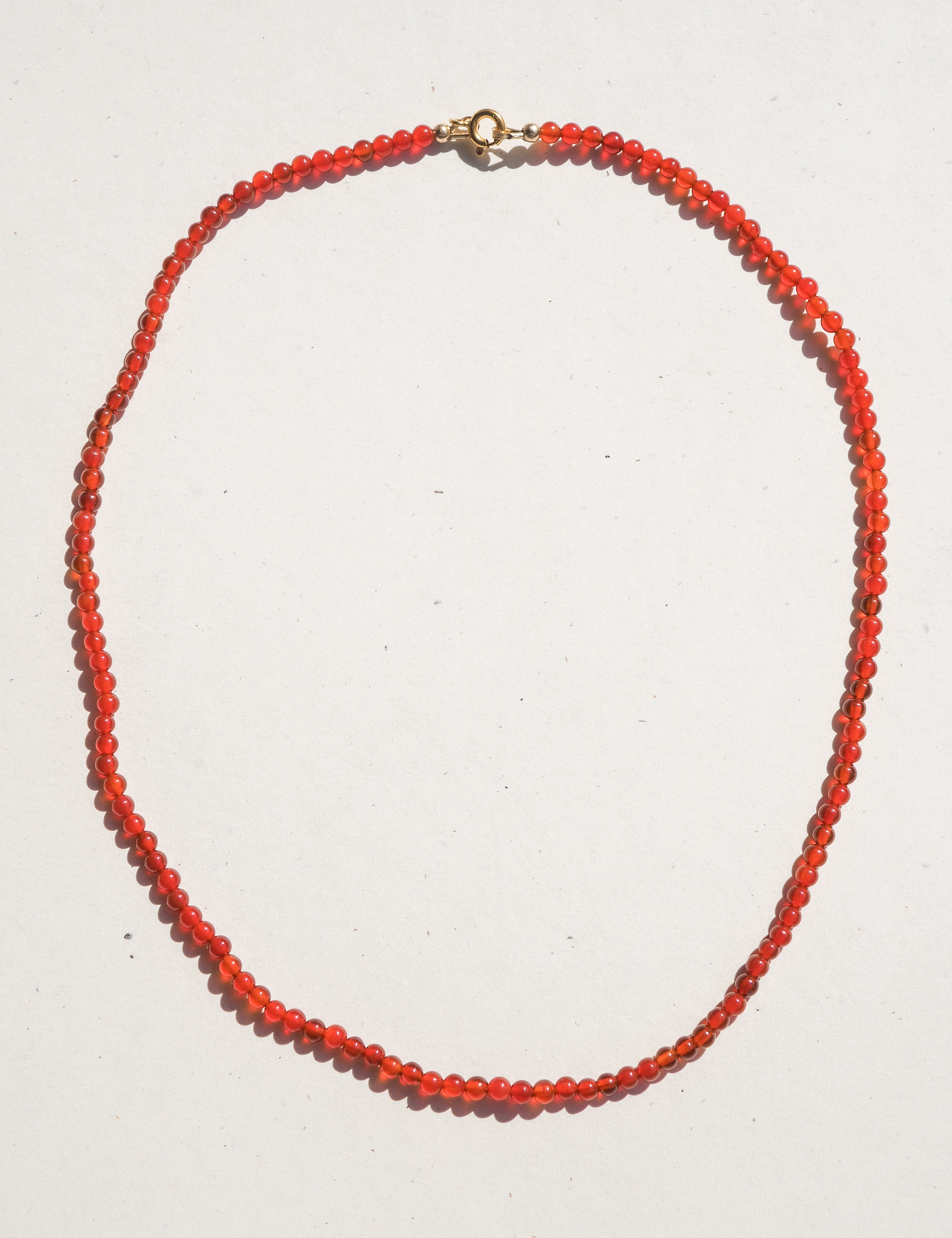 A beaded necklace made of small, faceted orange-red beads with a gold clasp, laid out in an oval shape on a plain white background.