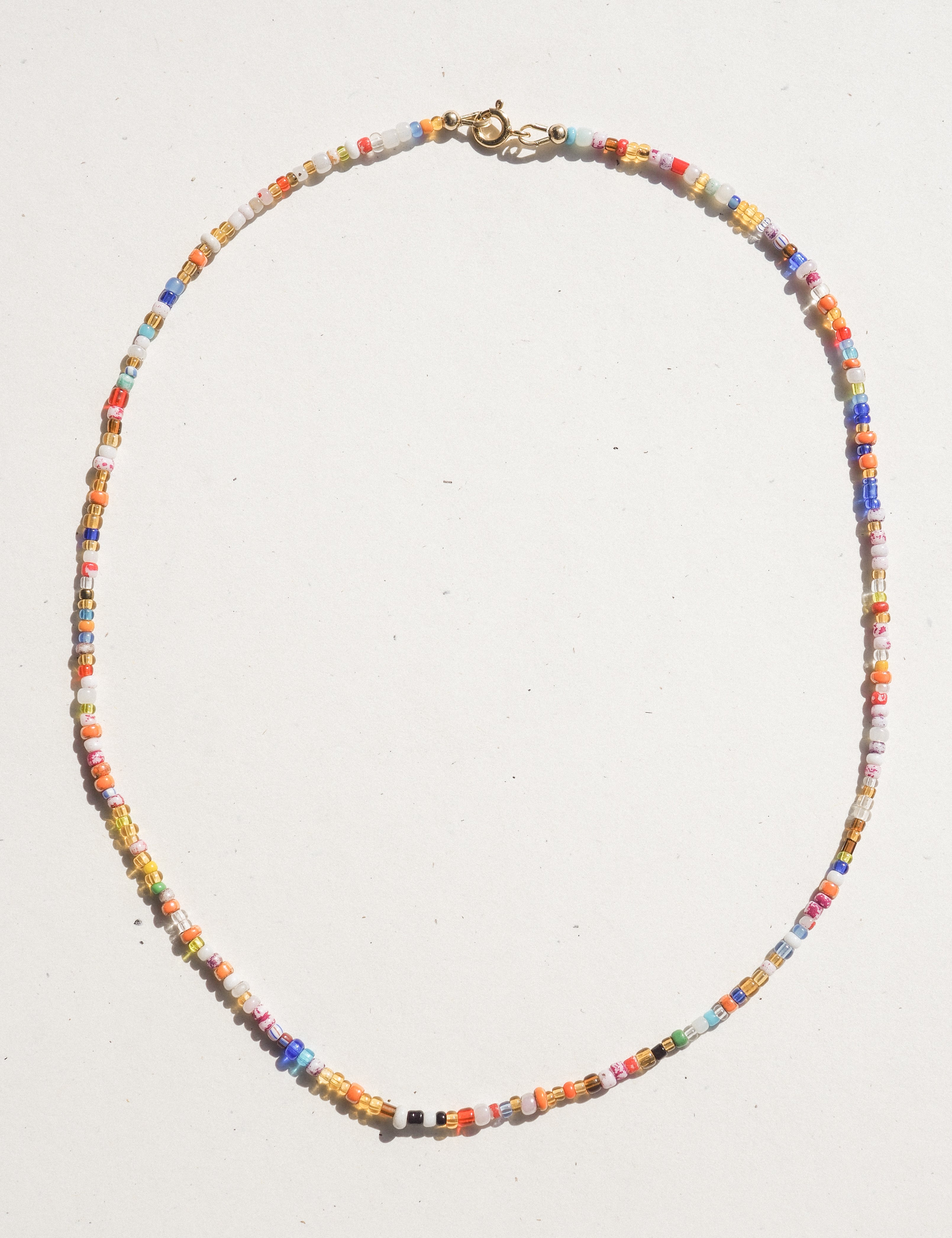 A delicate beaded necklace made of small multicolored cylindrical beads, arranged in an oval shape on a plain off-white background. It features a gold-toned lobster clasp at the top.