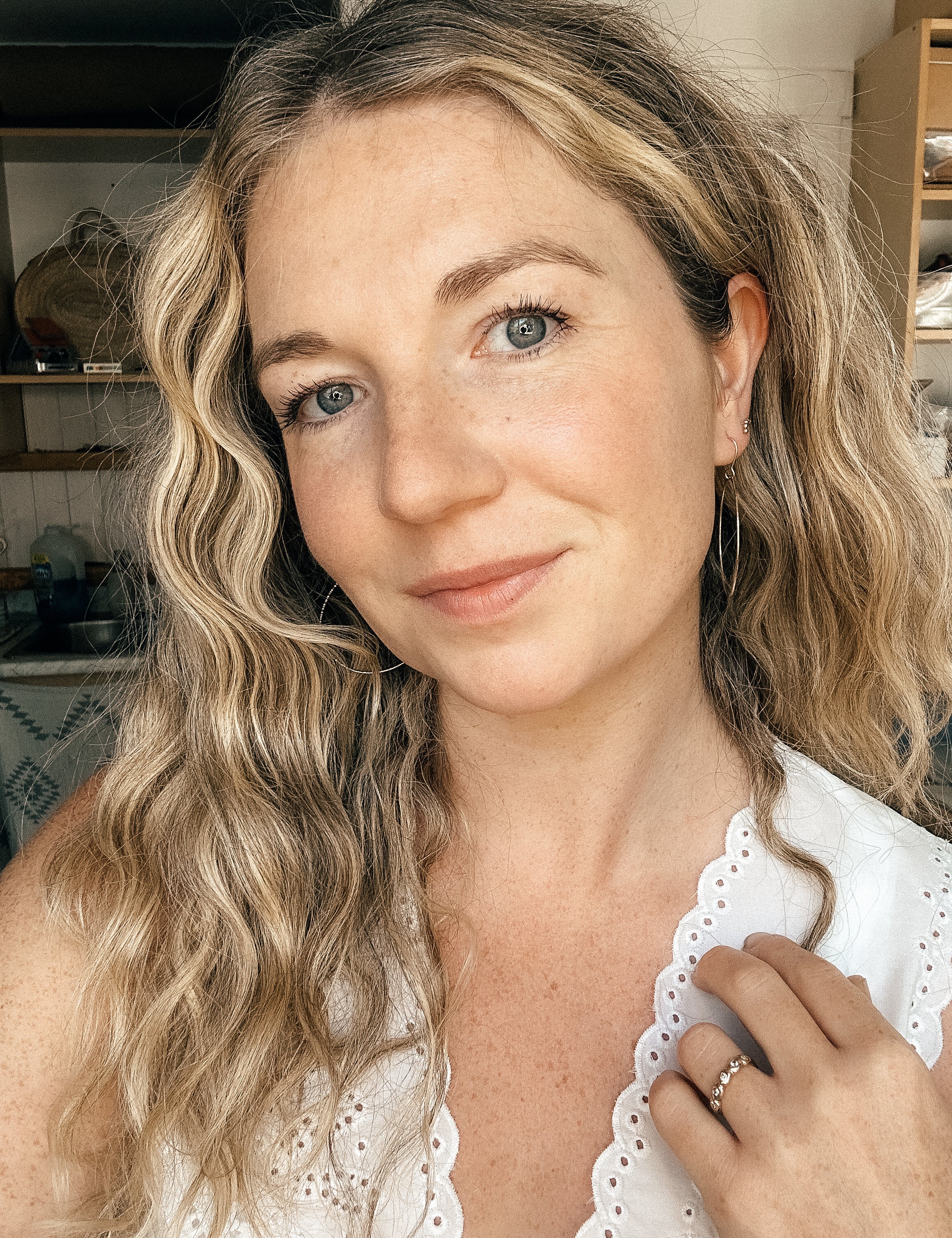 A close-up portrait of a woman with wavy blonde hair wearing a white eyelet lace top, with a ring on her finger, set against an indoor background.
