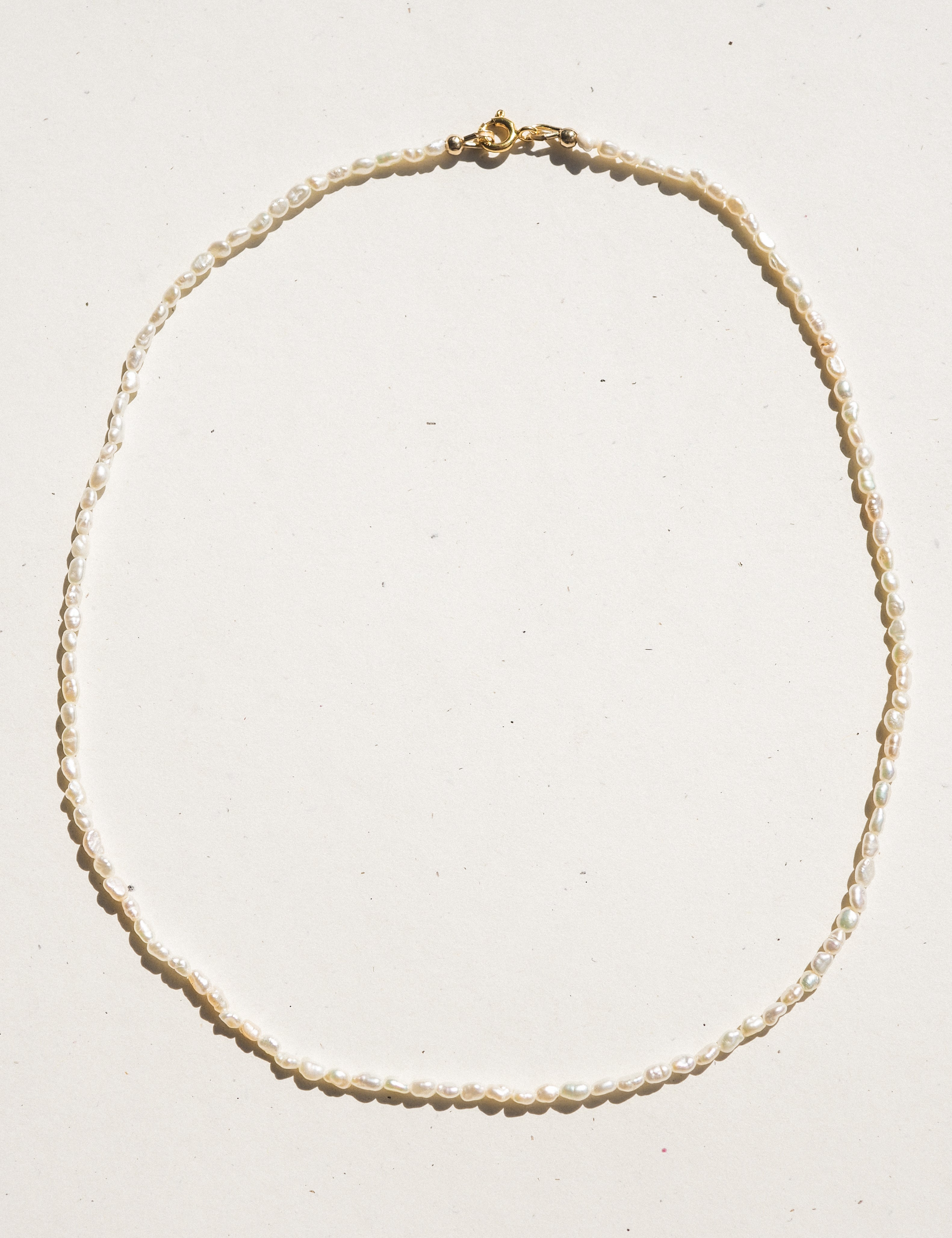 A delicate beaded necklace made of small cream-colored pearls with a gold clasp, laid flat on a light off-white background.